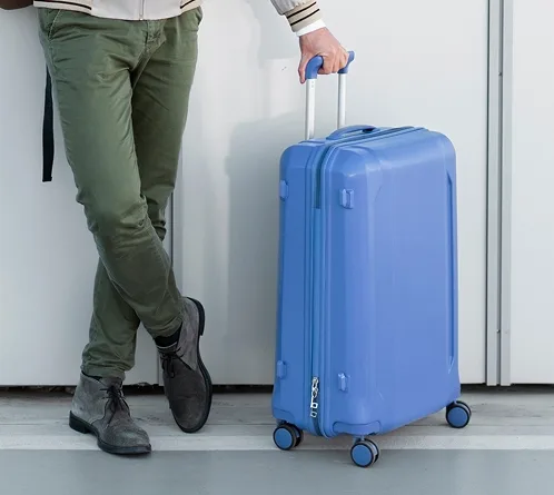 Persona de pie junto a una maleta rígida azul, sosteniendo el asa